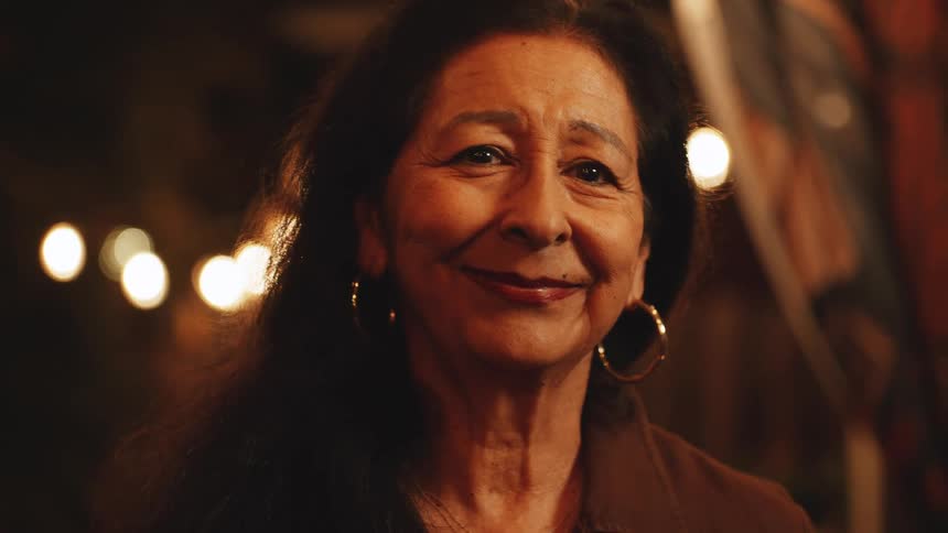 In a warm intimate close up an elderly woman with a kind and knowing smile looks directly at the camera