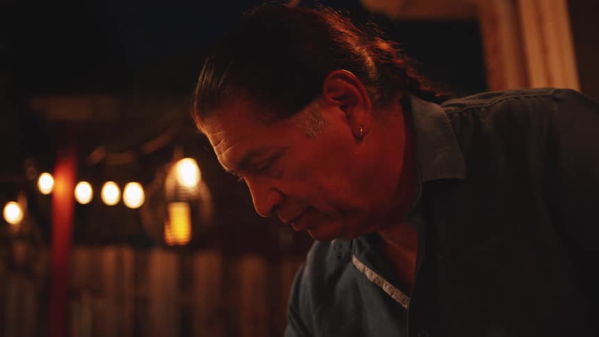The video is a closeup of a middleaged man with dark hair tied back bending over and focusing on something below him he is lit by warm string lights hanging in the background suggesting an evening or nighttime outdoor setting
