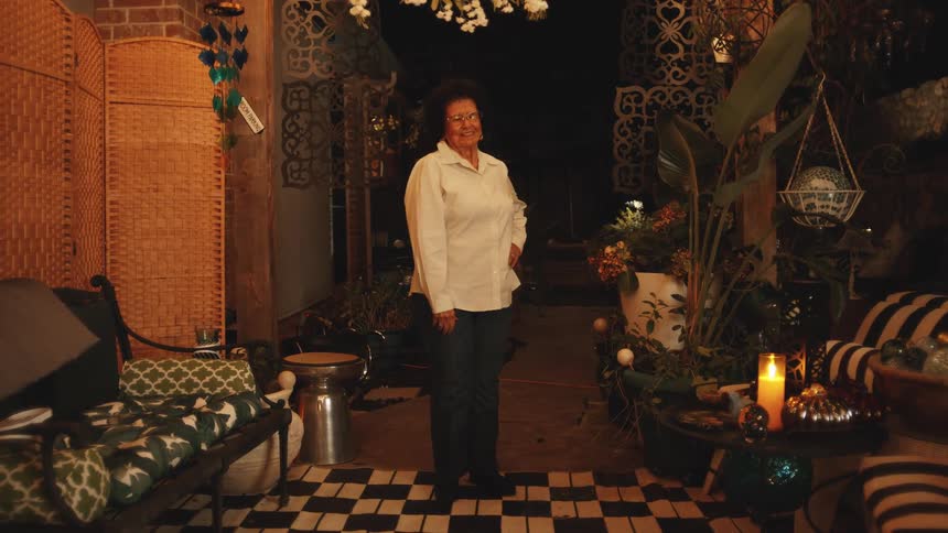 A smiling elderly woman stands proudly with her hand on her hip in the middle of a beautifully decorated warmly lit patio at night