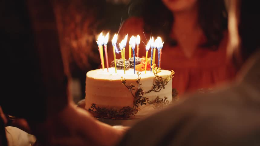 The video is a closeup of a celebratory moment with a birthday cake adorned with several lit candles of various colors two people are holding the cake while a third older person smiles brightly in the background