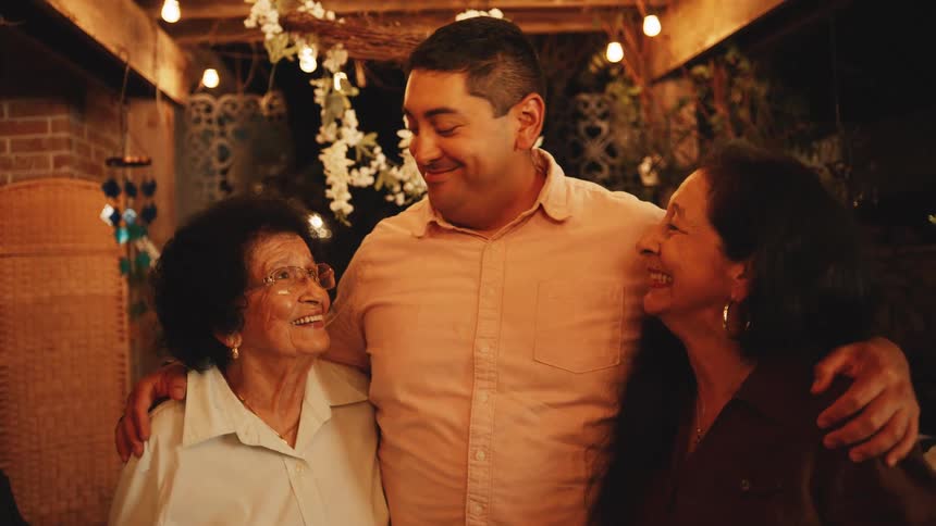 A middleaged man stands on the front porch at night holding close two womenone middleaged and the other elderlyas they all smile warmly bathed in the soft glow of porch lights