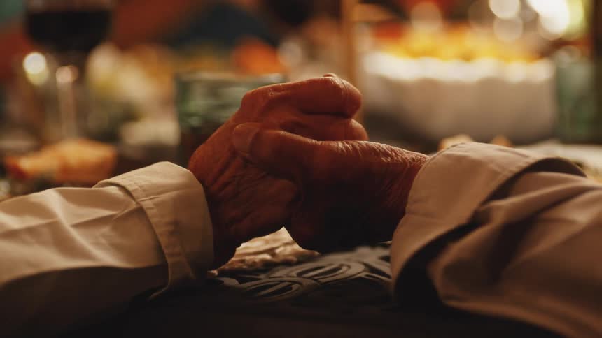 The video is a closeup of a persons older wrinkled hands clasped together on a table possibly in prayer or deep thought at a dinner gathering the background is softly blurred but shows wine glasses plates and a cake indicating a warmly lit