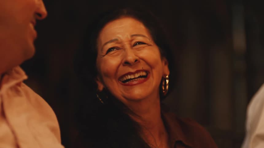 The video is an extreme closeup of an older woman smiling and talking to someone just out of frame she has dark hair and is wearing gold hoop earrings and a simple necklace