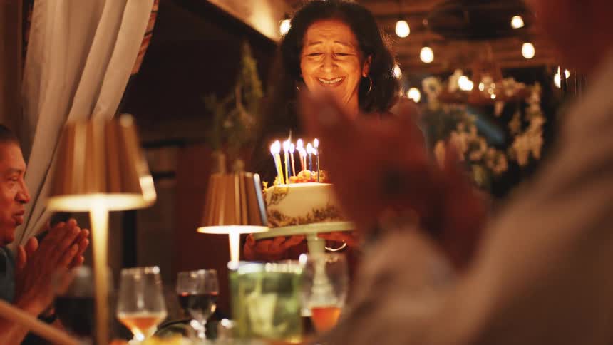 Older woman smiles while presenting a birthday cake as others sitting at the table clap