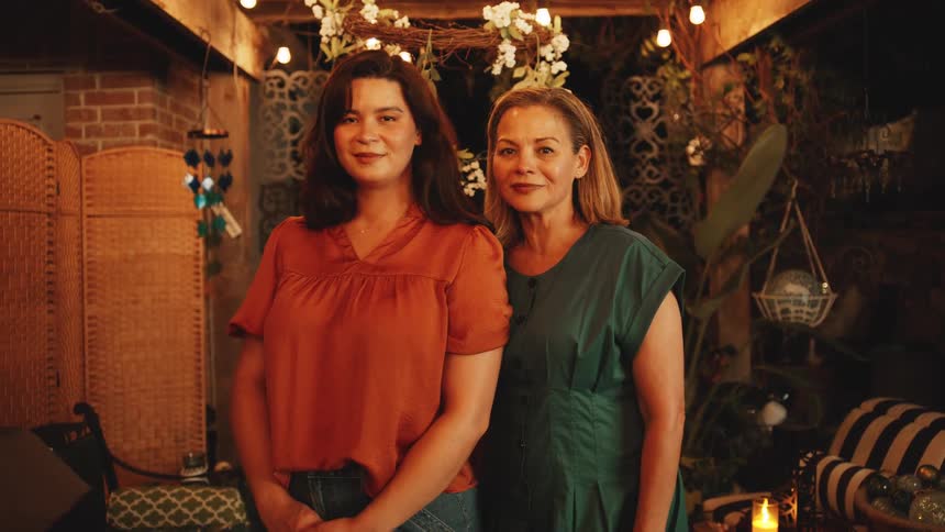 The video is a static medium shot of two women standing sidebyside in a warmly lit bohemianstyle outdoor or covered patio space the setting is decorated with string lights greenery and a candle creating a cozy evening atmosphere