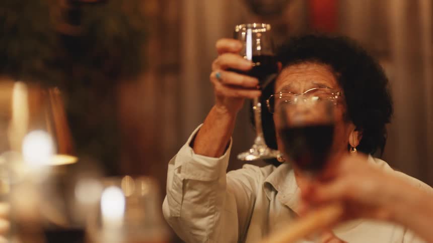 An older woman seated at a dinner table raises a glass of wine high she smiles and speaks to people across the table other people raise their glasses to meet hers and the scene is warmly lit with ambient indoor lighting