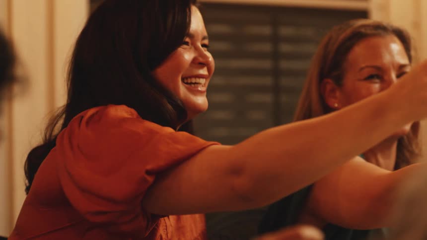 A woman with dark hair wearing an orange shirt raises a glass of red wine smiles laughs and looks around at other people during a gathering