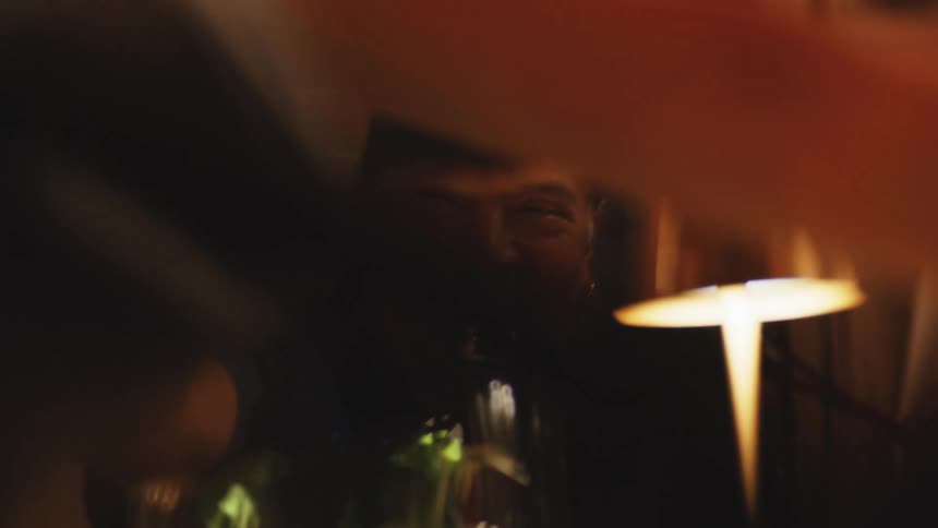 A man at a table smiles and laughs directly at the camera while raising a glass of red wine with the view partially obscured by blurred persons