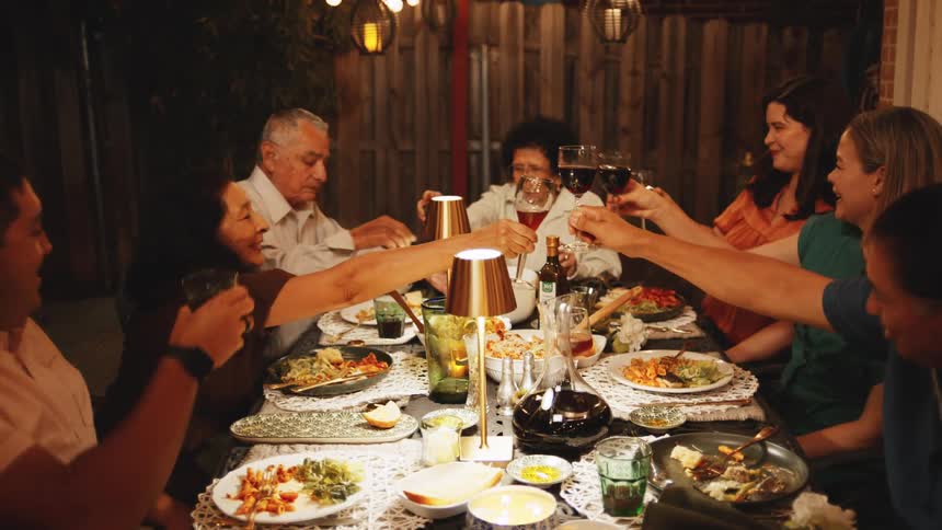 A group of seven people sit around a long dinner table under warm string lights at night raising their glasses to make a toast during the meal