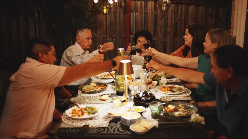 The video shows a group of seven people gathered around a large outdoor table at night raising their glasses to make a celebratory toast during a dinner the table is set with multiple dishes drinks and is lit by warm hanging lights and a centr