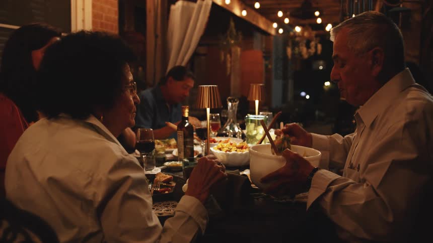 A group of diverse friends or family members are sitting around a table eating dinner and talking outdoors at night illuminated by string lights and a small gold table lamp as an older man in a white shirt serves himself from a large white bowl