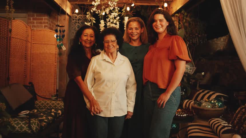 A medium view shows four smiling women of different ages posing together for a family portrait in a cozy dimly lit outdoor patio decorated with string lights and white flowers
