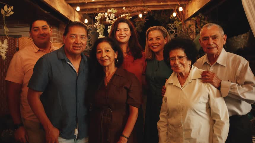 Seven people of various ages stand close together in two rows smiling and posing for a group portrait under a wooden patio structure decorated with string lights and white flowers at night
