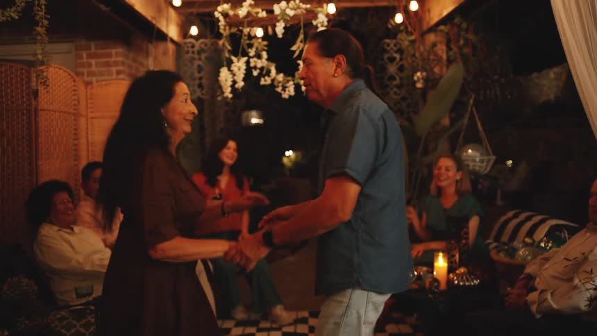 An older man and woman hold hands and dance in a dimly lit decorated patio space surrounded by seated people spinning and laughing before the man dips the woman in a final flourish