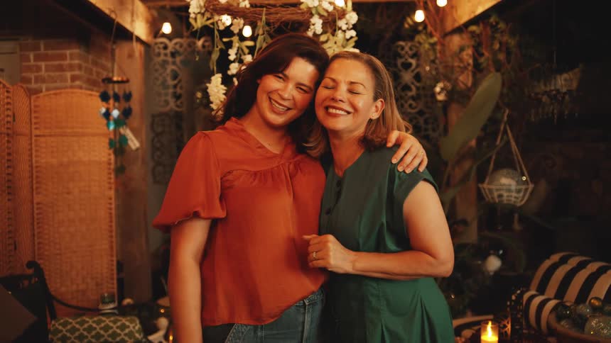 Two women standing close together under a covered outdoor patio with string lights initially talking and touching faces then laughing and embracing one another warmly