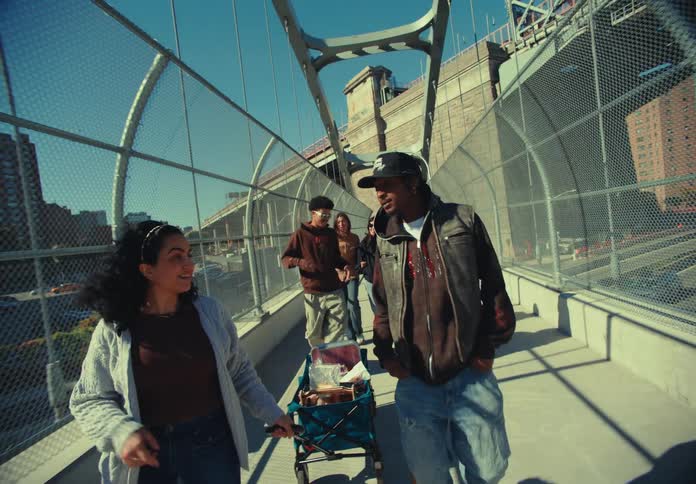 A camera tracks a group of people walking and interacting on a pedestrian bridge over a busy highway on a sunny day