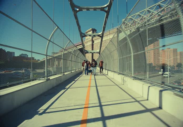 A group of people walks toward the camera along a pedestrian walkway on an elevated bridge bordered by metal support structures and wire fences with tall apartment buildings visible in the distance under a blue sky