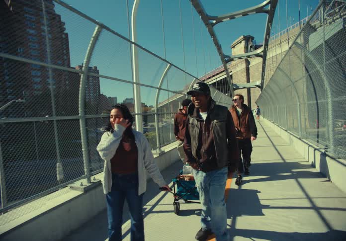 A group of people walks along a bridge walkway one person pulls a cart tall fences line the walkway buildings are visible in the background vehicle traffic moves beneath the path and the sun is shining