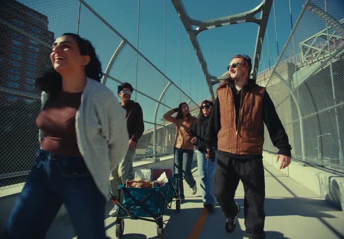 The video shows a group of young friends walking forward laughing and talking on a sunny pedestrian bridge that is enclosed by chainlink fences they are pulling a small wagon that appears to contain drinks and snacks for an outing