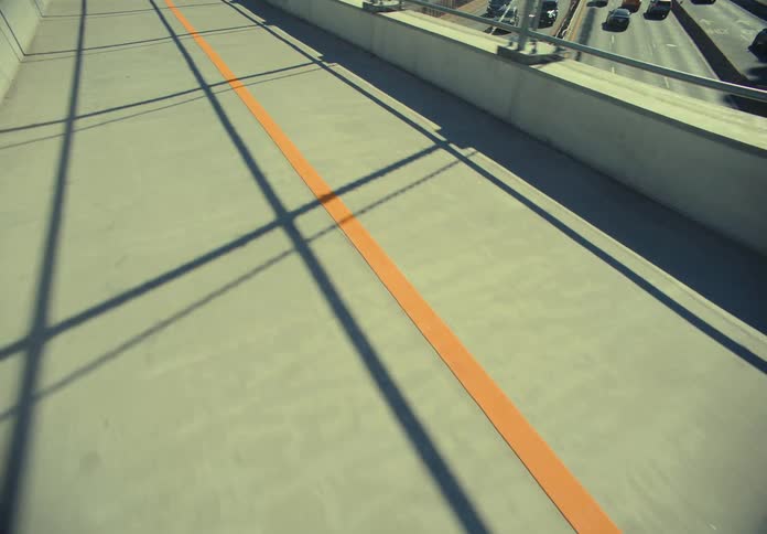 Orange line stretches along a sunlit walkway above a busy road