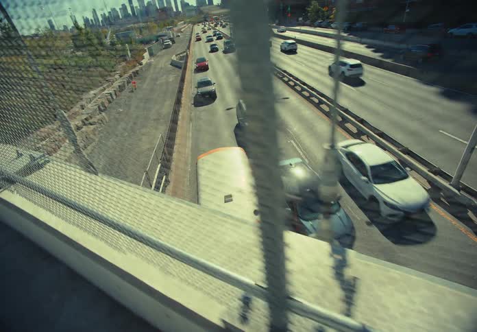 A busy city highway stretches beneath an overhead bridge on a sunny day with vehicles streaming past in both directions and sunlight glinting off the concrete and metal structures