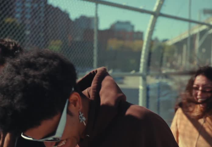 Three people walk past the camera on an outdoor pedestrian walkway with a chainlink fence smiling and laughing in the sunlight as city buildings are visible in the background