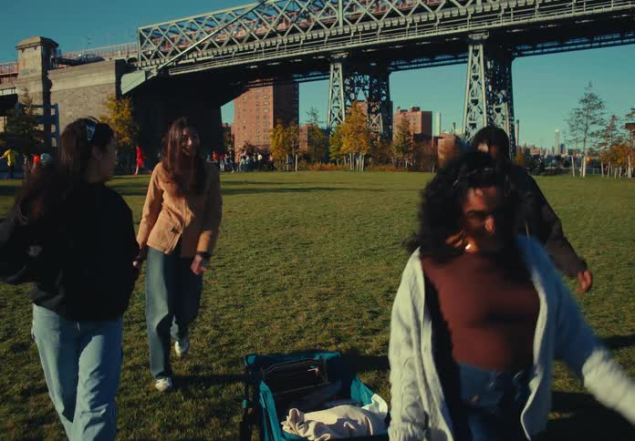 A group of four people including three women and one man walk forward across a grassy park while pulling a small blue wagon with a large steel bridge and brick buildings visible under a sunny blue sky in the background