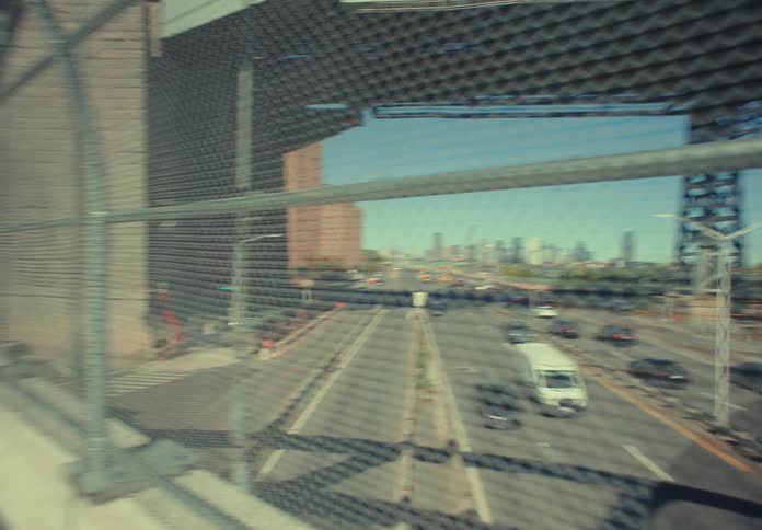 The video shows an undercarriage view of a massive suspension bridge from a moving vehicle on a highway with the lower bridge deck and steel truss work dominating the frame through the chainlink fence on the left a new york city skyline is