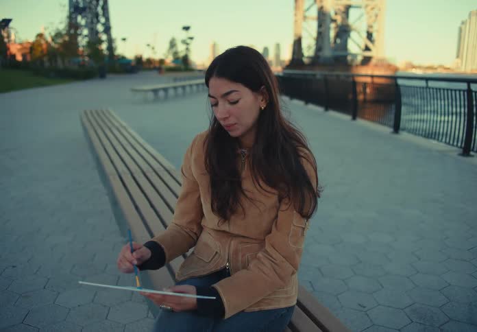 A young woman wearing a jacket sits on a public bench at midday using a pen to make strokes on a piece of cardboard resting on her arm focused and absorbed in her task
