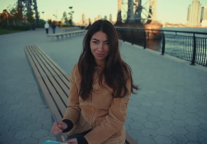 A woman with long dark hair sits on a wooden bench in a paved waterfront park looking down as she applies blue paint to a canvas then pauses looks up and smiles toward the camera with the city skyline and a bridge visible behind her