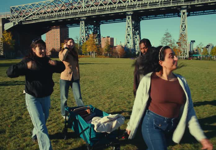 Four people walk together across a sunny grass field two women on the left and a man and a woman on the right the woman on the right pulls a blue folding wagon and a large metal bridge and city buildings are visible in the background