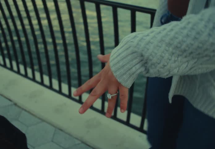 A young woman inserts a ring into her partners finger while standing on a bridge at sunset