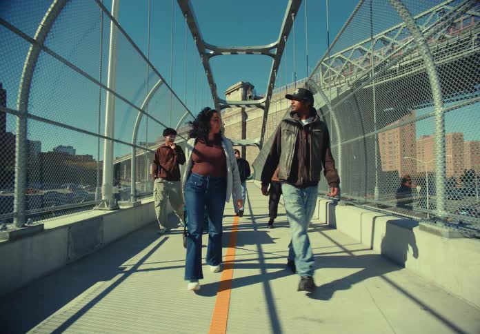 A group of young people is shown walking toward the camera on a pedestrian walkway of a bridge under a clear sunny sky the walkway features chainlink fences on both sides and a painted orange and white line running down the center with