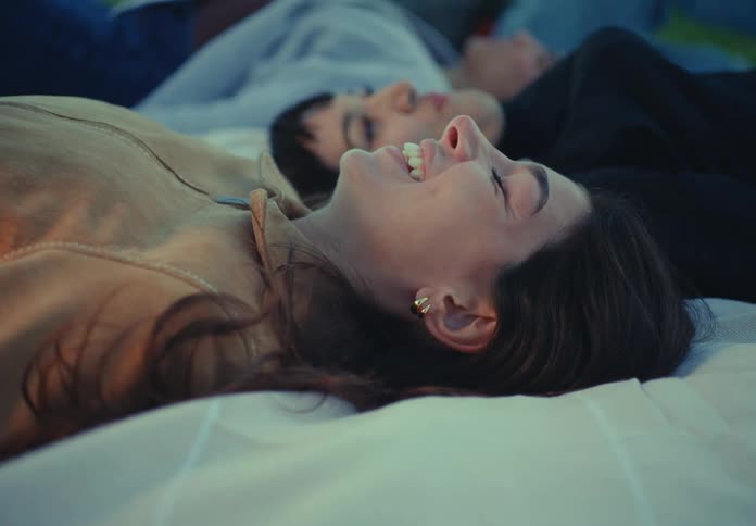 The video is a closeup of a person lying down on a blanket with their friends in a park setting they are laughing and smiling brightly while looking up toward the sky