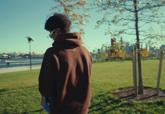 A man with an afro hairstyle and sunglasses wearing a brown hoodie and khaki pants removes a folding game board from a turquoise bag on the grass and then sets it up in a sunny park with a bridge and city skyline in the background