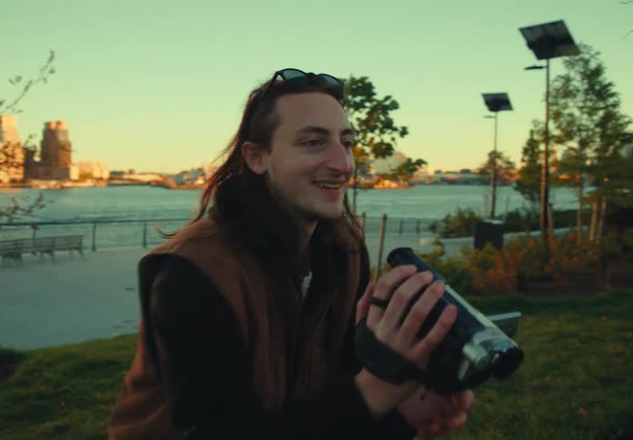 A man with long hair wearing sunglasses on his head stands on a grassy hill in an urban park smiling while holding a video camera with a large suspension bridge and city skyline visible across the water in the background
