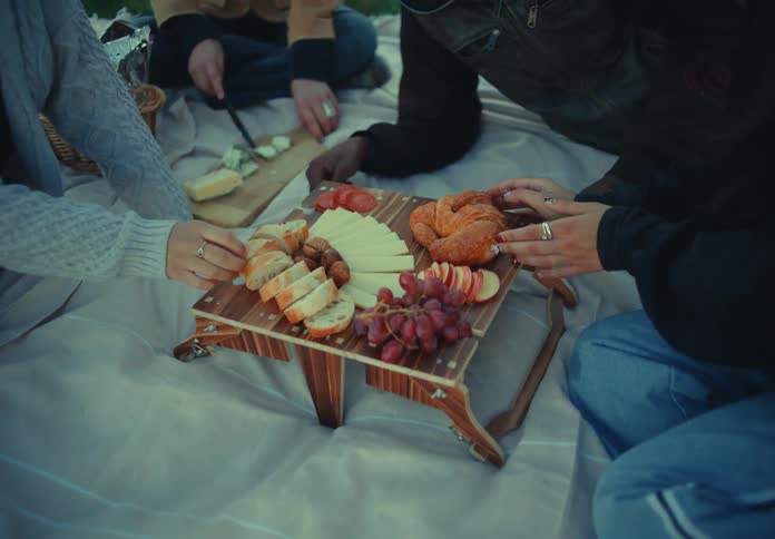 The camera moves down from green grass to a blanket a group of people sits on the blanket a low wooden platter holds food including cheese and grapes peoples hands reach toward the food and knives and other food items rest nearby