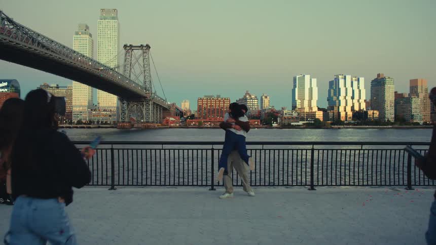 A man and a woman stand by a waterfront railing in a city park at sunset where they embrace and kiss while a group of people suddenly appear from the sides and shoot multicolored confetti into the air