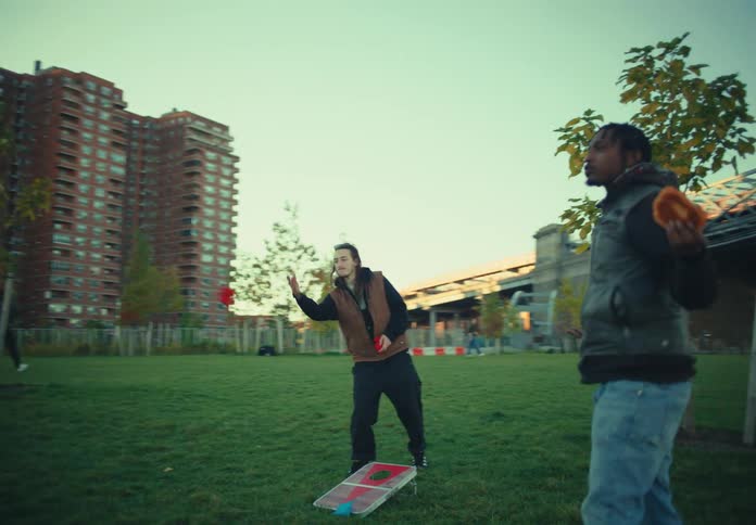 A group of people enjoys a picnic with food on a blanket in a park with grass bordered by tall apartment buildings and a steel bridge with some individuals playing a beanbag toss game