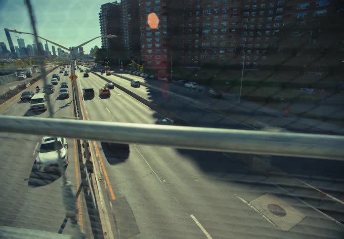 An overhead bridge spans a busy city highway on a sunny day with vehicles streaming below and sunlight glinting off the urban structures