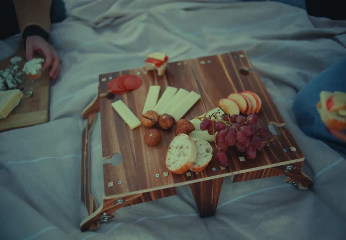The video is a closeup of a wooden portable picnic table set on a blanket laden with a variety of snacks including cheese grapes croissant sliced bread and hazelnuts multiple hands reach into the frame to pick up the food from the platter
