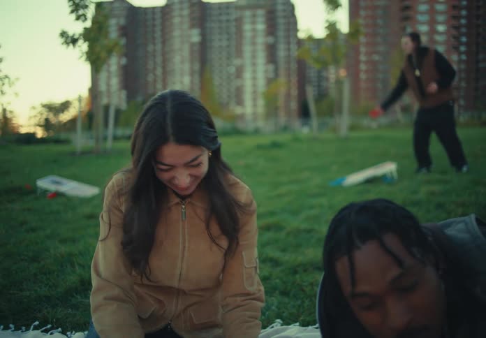 The video is a medium closeup of a young woman sitting on a blanket in a park who is smiling and laughing joyfully while looking at friends who are out of frame the background features tall apartment or office buildings in an urban park setting