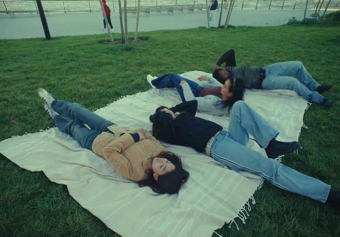 The video begins with a lightcolored blanket being laid out on a grassy field near a paved waterfront path a group of four friends then lies down on the blanket sidebyside relaxing and enjoying the park setting