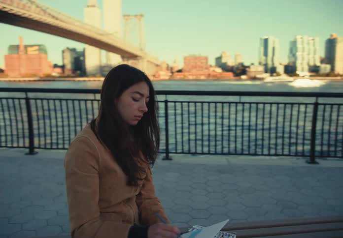 A woman sits on a long wooden bench at a paved waterfront park painting a canvas and looking out at the city skyline