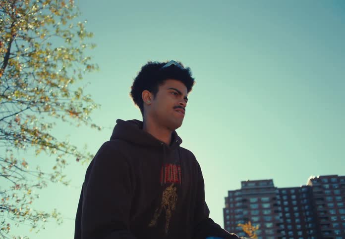 A man in a dark hoodie stands outdoors holding blue bean bags and throws one bag with a smooth motion toward the right with apartment buildings and trees visible in the background under a bright clear sky
