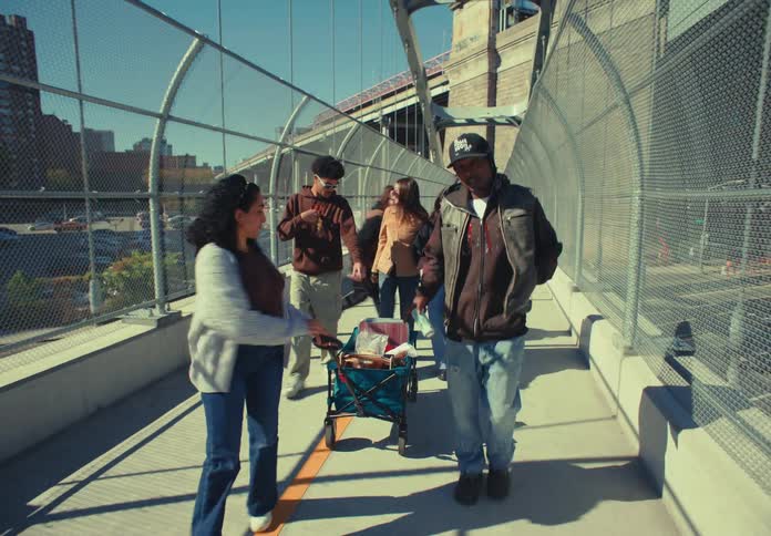 A diverse group of friends in casual clothes walks along a pedestrian walkway with wire fencing smiling and carrying a small folding cart on a sunny day