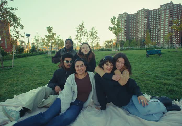 Five friends walk across a sunlit grassland at midday then settle onto a blanket in the park smiling as they prepare for a cheerful picnic
