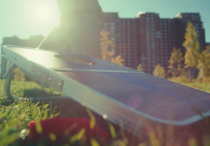 A lowangle shot shows a board pitched on a sunny grassy lawn in an urban park where a person stands and throws a red bean bag that bounces off the board with tall apartment buildings and yellowleafed trees visible in the bright background