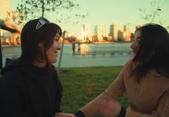 Two female friends sit on a grassy stretch in a public park deep in conversation under the midday sun suddenly one of them stands up with a burst of energy and runs toward the nearby pathway her friend watching with a smile as the moment unfolds
