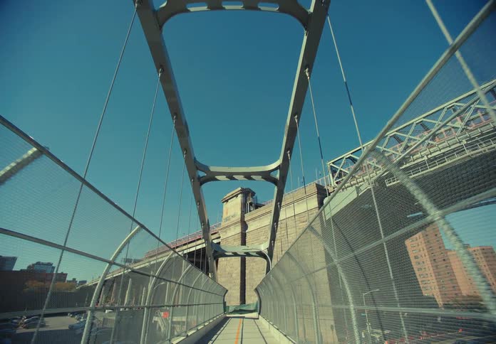 A camera moves steadily along a fenced elevated pedestrian path on a large steel bridge passing under metal supports toward a stone tower structure with traffic and city buildings visible on both sides under a clear blue sky